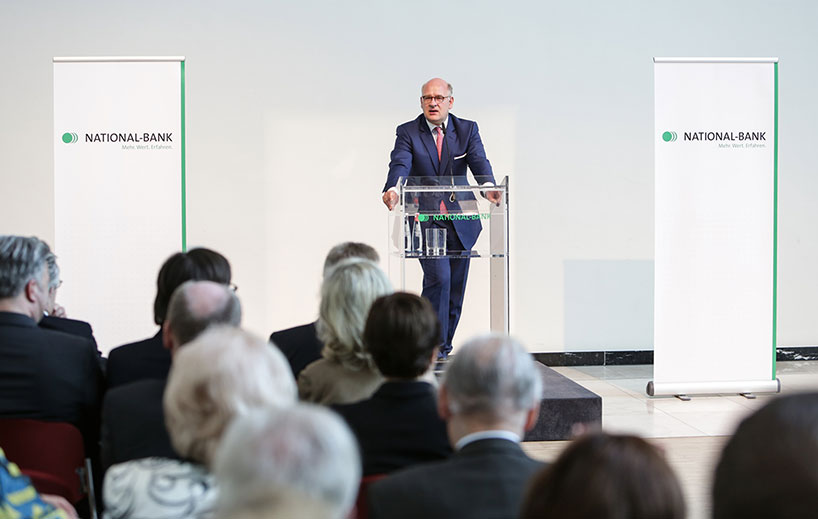 Hr. Dr. Lange am Rednerpult flankiert von zwei Rollups mit National-Bank-Logo. Zuhoerer unscharf im Vordergrund.
